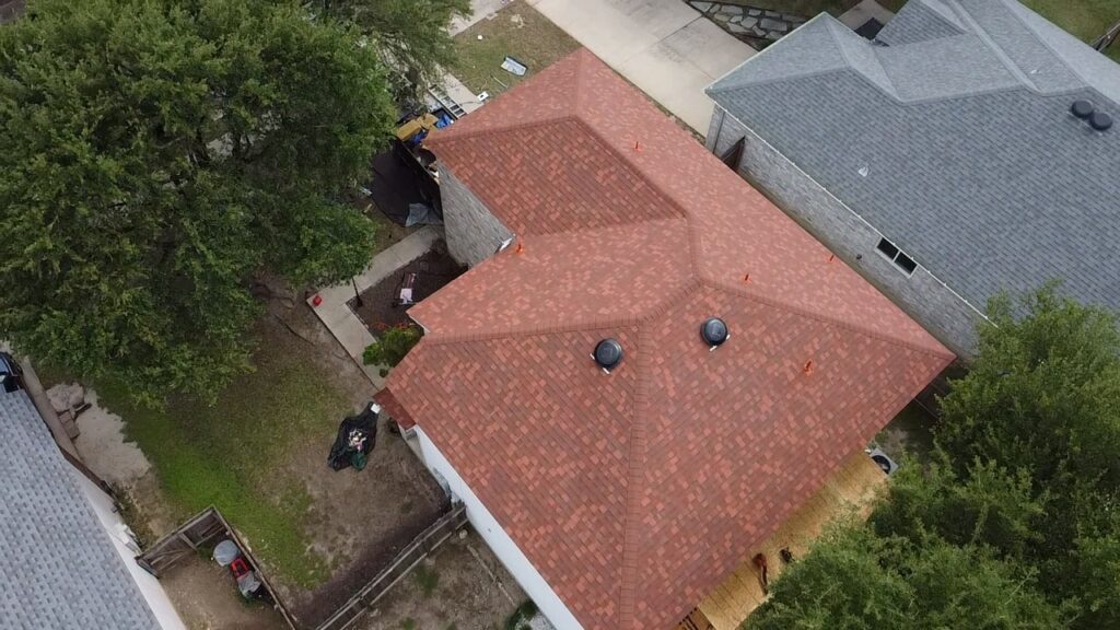 Overhead view of a reddish shingle roof with intersecting roof planes beside mature trees