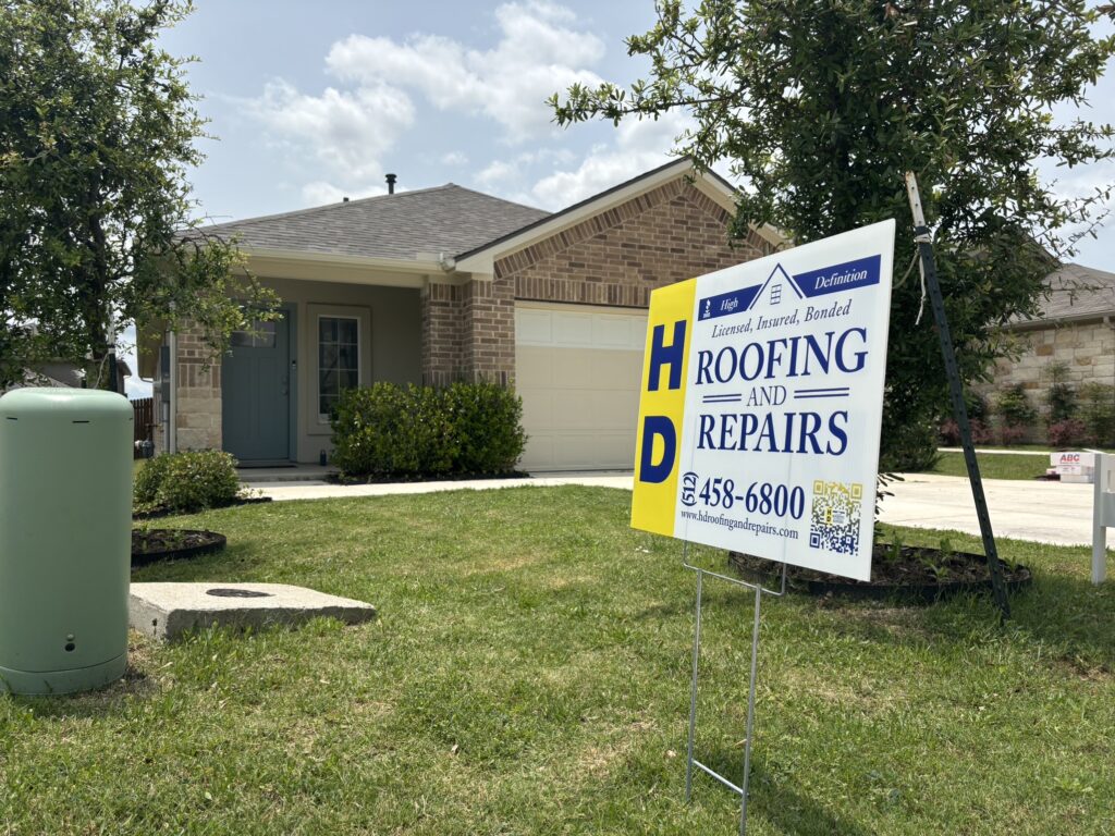 A shingle-covered roof on a brick home with an HD Roofing sign in the yard
